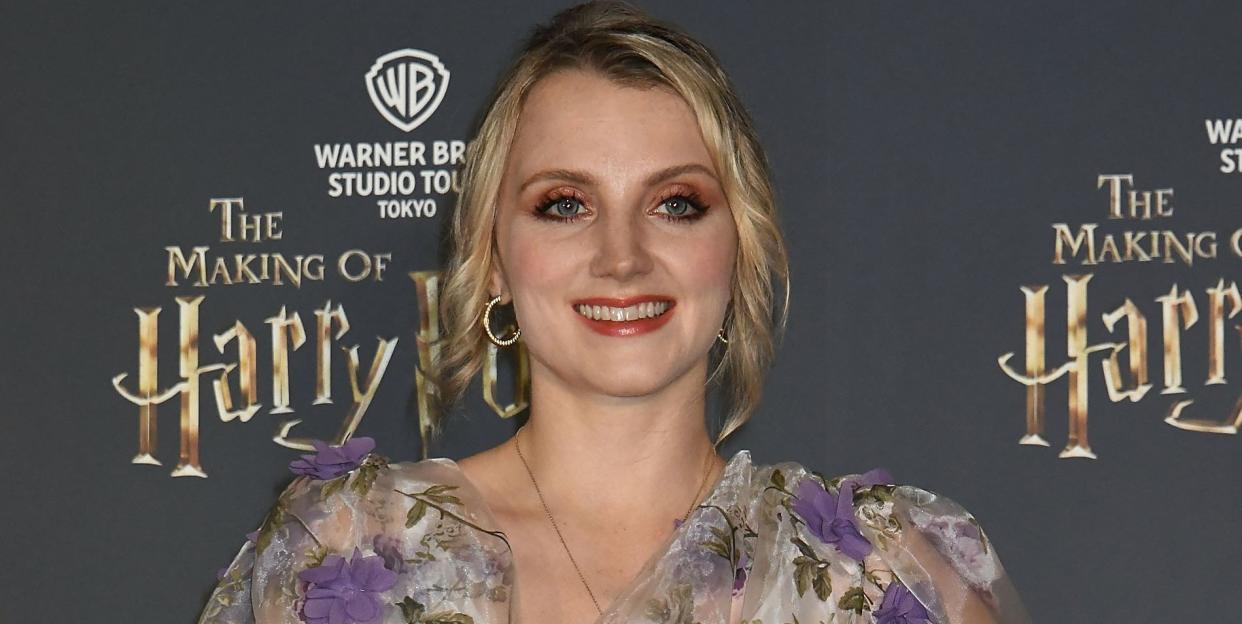 evanna lynch, a young woman stands looking at the camera with a smile and her left hand on hip, she has blonde hair worn up with curls loose framing her face and wears a white dress with purple flowers, she hold a paper fan open in her right hand