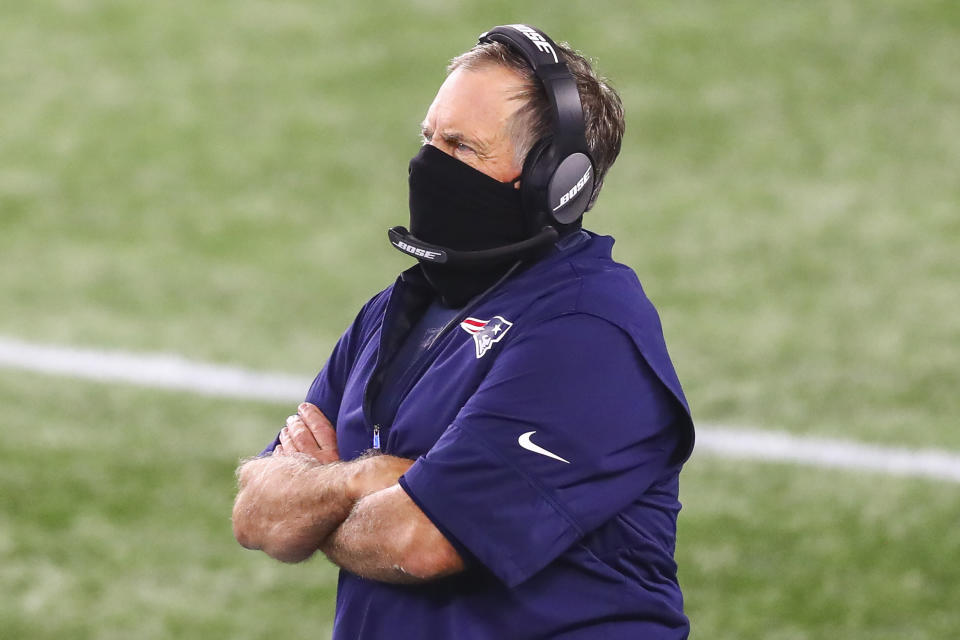 Bill Belichick wears a mask and a headset with his arms crossed.