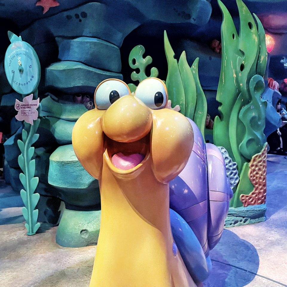 A cartoon snail character, Gary, from SpongeBob SquarePants, is smiling with an open mouth. He is in an underwater-themed attraction with seaweed and rocks