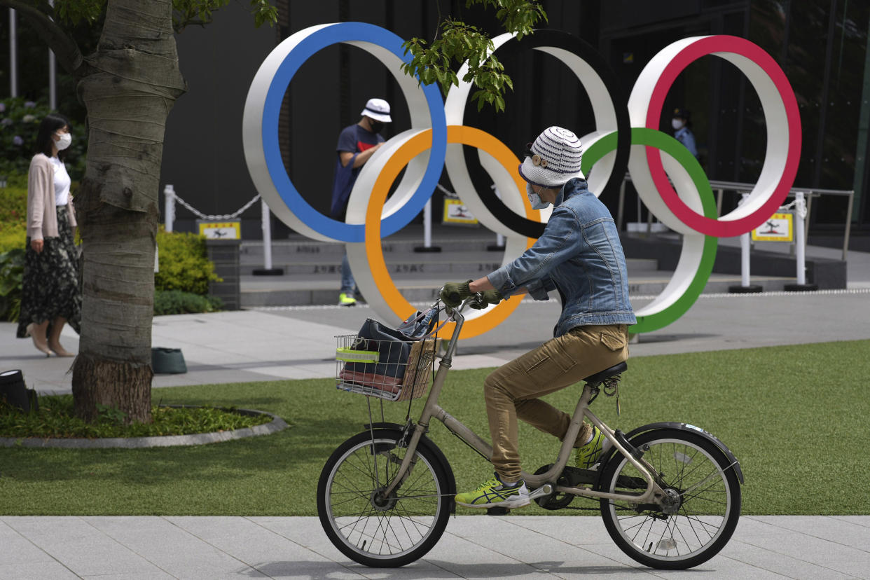 Image: A woman rides a bicycle near the Olympic Rings Wednesday, June 2, 2021, in Tokyo. (Eugene Hoshiko / AP)