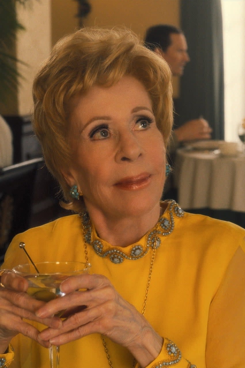 Carol Burnett sits at a restaurant table holding a drink, wearing an elegant, embellished yellow outfit. Other diners are engaged in conversation in the background