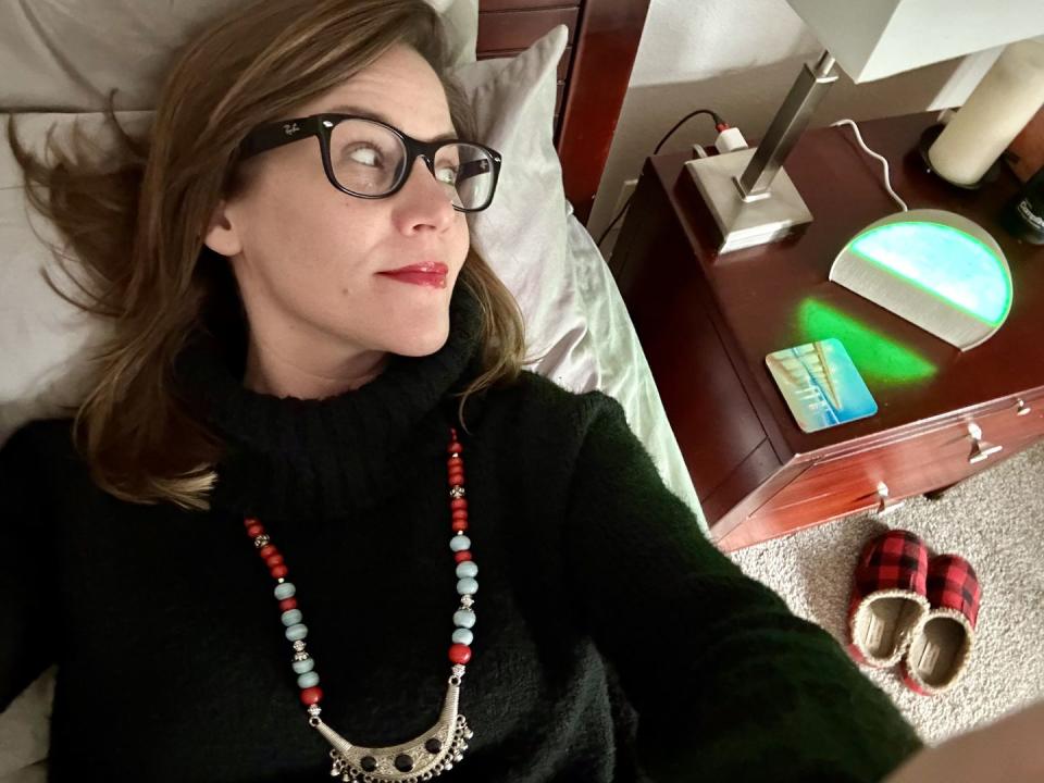 a woman wearing glasses lying on the bed, staring at her alarm clock