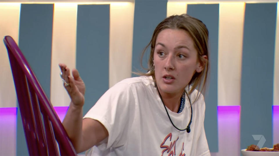Estelle in an argument with her hand up, wearing a white t-shirt and hair pulled back in the Big Brother house