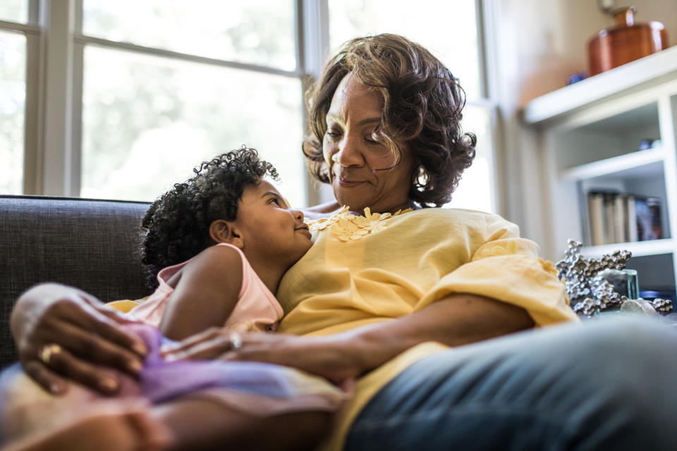 An elderly woman lovingly embraces a young child on a sofa in a living room setting. Both appear content and close. 