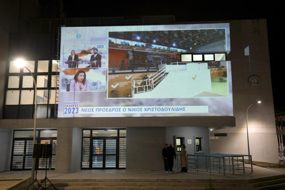 People stand under a giant screen reporting that Nikos Christodoulides has been elected as the new President, outside of a stadium where Christodoulides will arrive for his inauguration ceremony, in Nicosia, Cyprus, Sunday, Feb. 12, 2023. Former foreign minister Nikos Christodoulides has been elected as the new president of Cyprus in a runoff election. (AP Photo/Petros Karadjias)