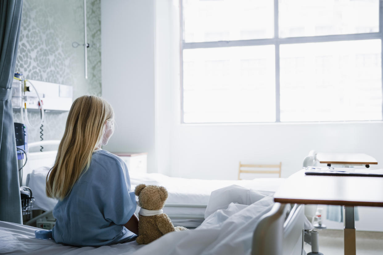 Little girl sitting on a hospital bed with her Teddy Bear by her side looking towards a window.