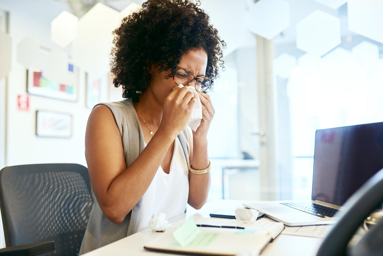 A woman holds a tissue to her nose as she sits at her office desk.