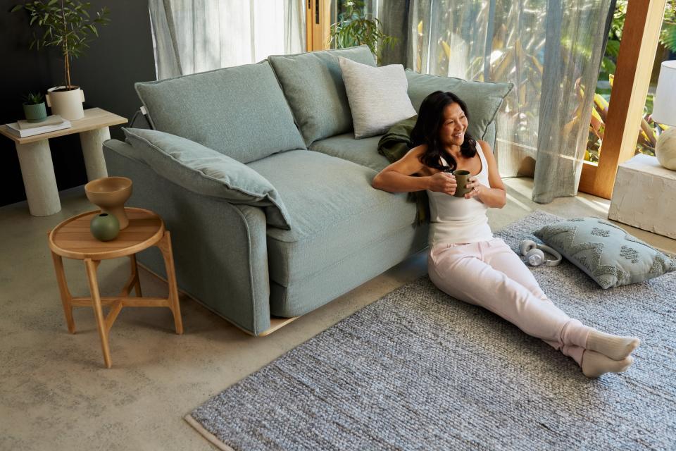 A model sits smiling on the ground leaning against a green Cushy Sofa Bed (Queen size) in a loungeroom setting.