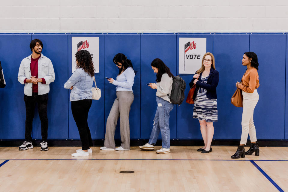 People are lined up against a gym wall with "VOTE" signs, waiting their turn. They are using their phones and talking