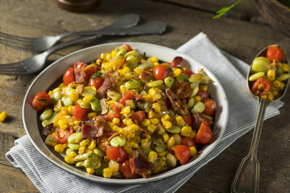 A bowl of succotash featuring corn, lima beans and pieces of bacon on a napkin with a spoonful on the side