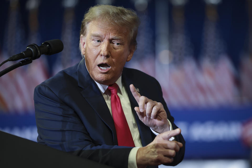 Donald Trump in a suit speaking at a podium with microphones, gesturing with his hands