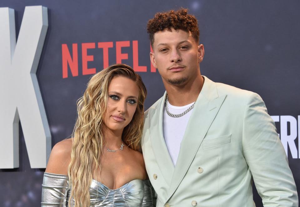 brittany mahomes and patrick mahomes pose for a photo in front of a dark gray background, she wears a strapless silver dress, he wears a baby blue suit jacket over a white shirt with a silver chain necklace