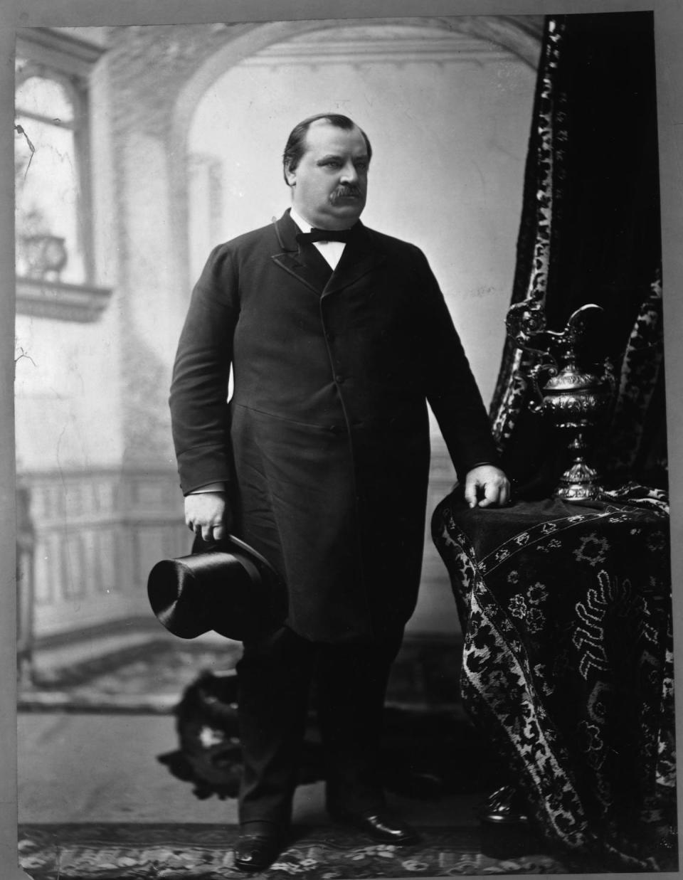 A suited man stands with a top hat in his right hand as his left hand rests on a side table dressed in a table cloth.