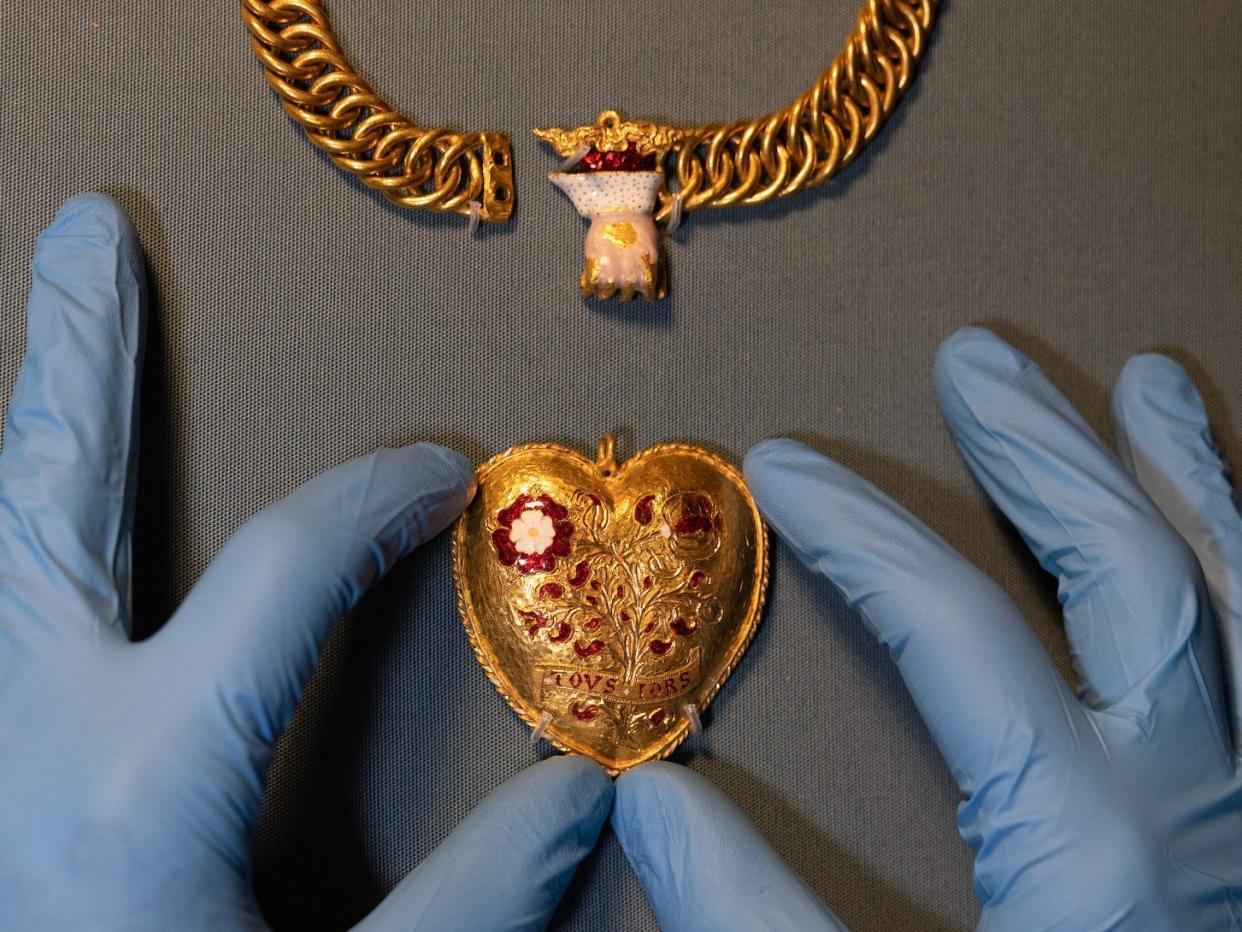 Two gloved hands hold the pendant, showing the front of the heart-shaped locket.