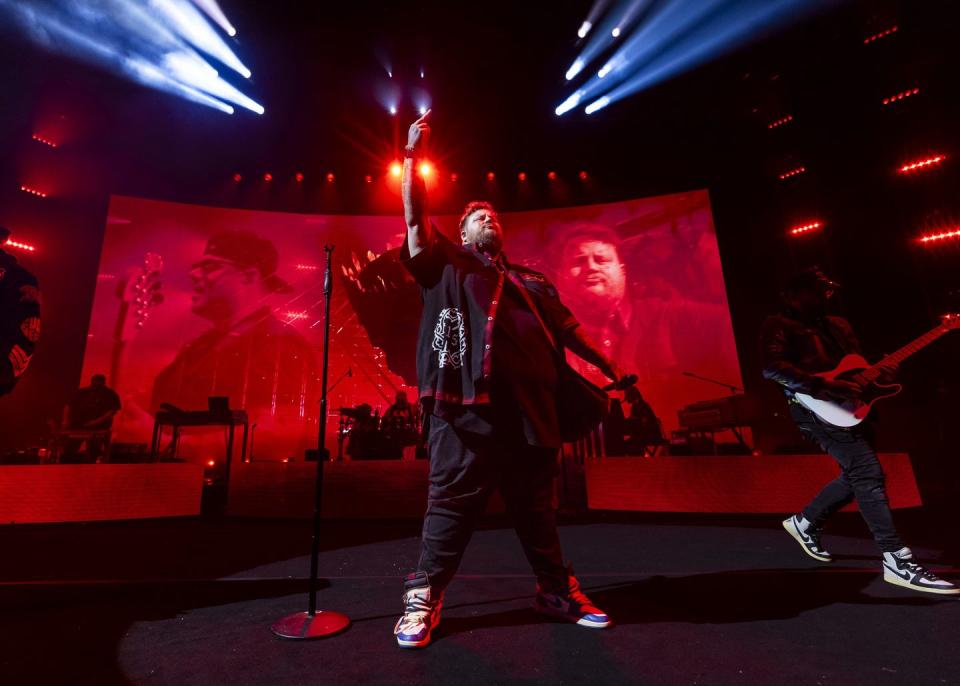 jelly roll stands on a stage with one hand pointing up in the air and another holding a microphone out at his side, he wears a black button up over a black shirt, dark pants, and sneakers, behind him is a large video screen and musicians with instruments