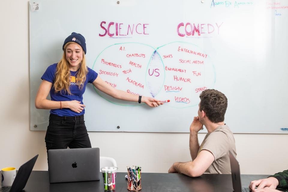 Sarah Rose Siskind in a blue t-shirt and beanie pointing to a white board with red writing during a meeting with work colleagues.