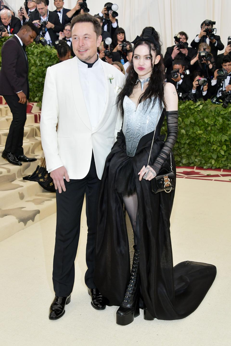 Elon Musk and Grimes standing together at the 2018 Met Gala. Musk is wearing a white blazer and Grimes is wearing a long dress with a black skirt.