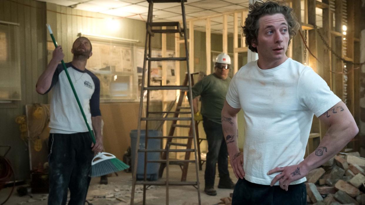  Ebon Moss-Bachrach looking at the ceiling while holding a broom and Jeremy Allen White standing with his hands on his hips. The Beef is torn a part behind them. 