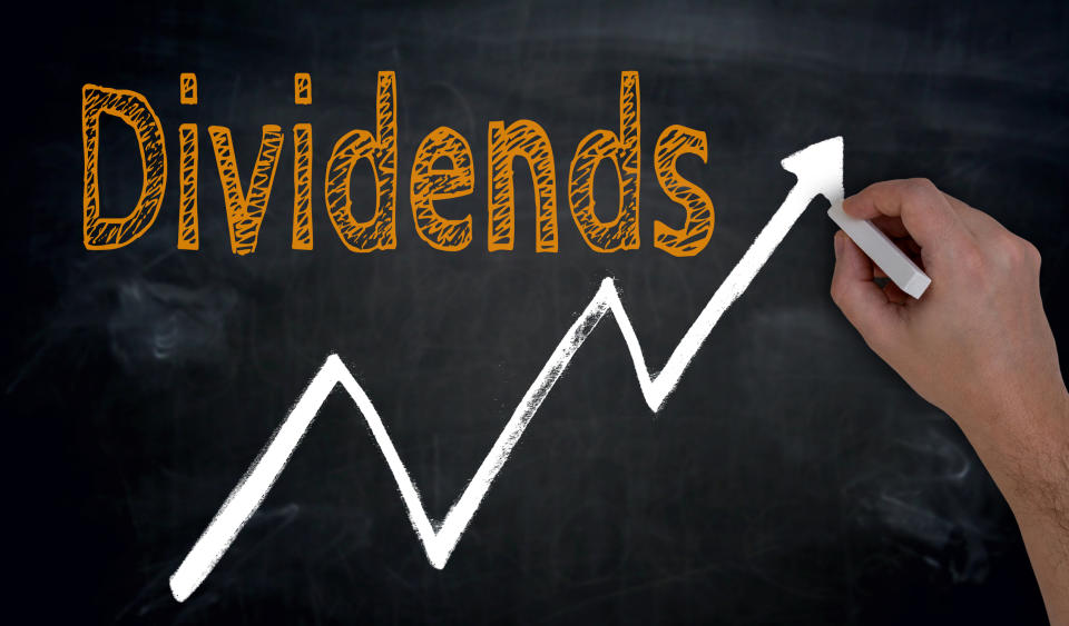 A hand draws an upward sloping line on a chalkboard with the word dividends in orange chalk above it.