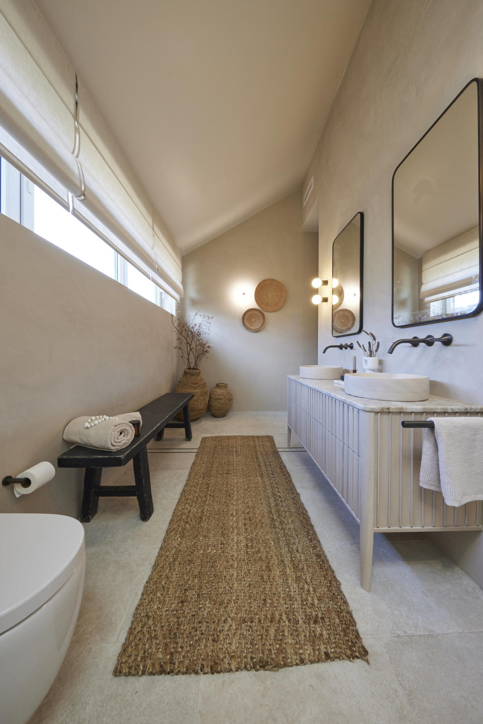 A wide shot of the bathroom with a bench on left, a vanity and mirrors on the right and a beige mat on the floor. 