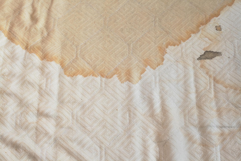 A fabric surface displaying a large, irregular stain and visible wear. The material has a patterned texture. No people or text are present in the image