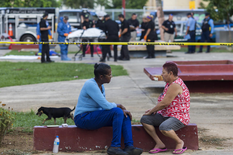 A couple of neighbors talk next to a strand of police tape as investigators in the background recover the bodies at the scene of a multiple killing in San Juan, Puerto Rico, Tuesday, Oct. 15, 2019. Several people are reported dead following a shooting in the Rio Piedras neighborhood of San Juan. (AP Photo/Dennis M. Rivera Pichardo)