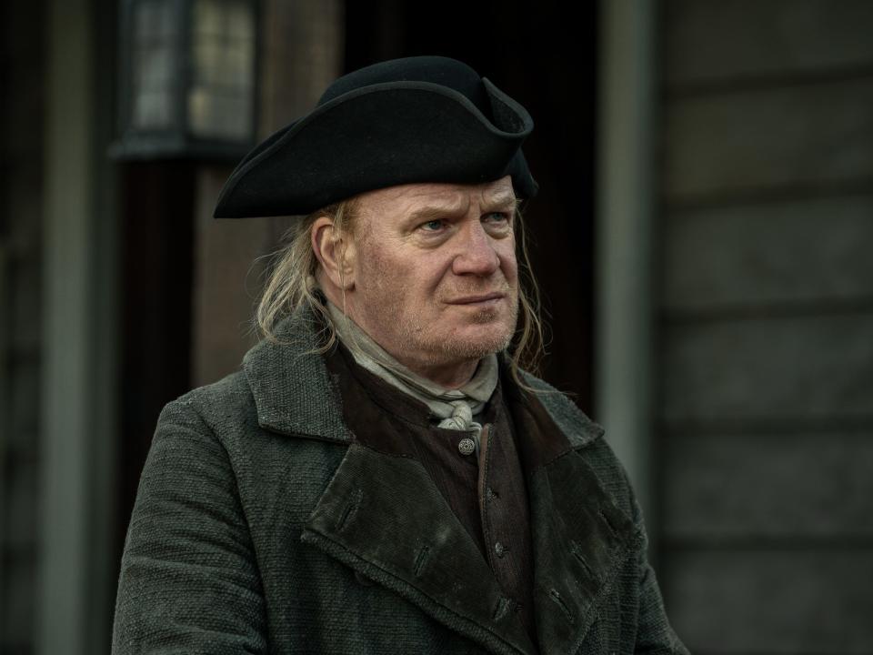 Tom Christie wearing a dirty-looking three-peaked hat and a grey overcoat while standing in front of a simple wooden home with a lantern hanging above the door.