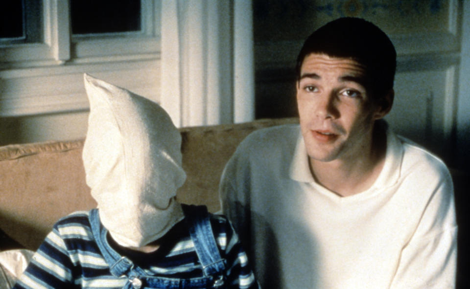 A young man sits on a couch, next to a child who has a bag over their head