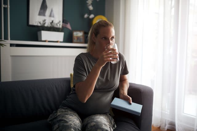<p>vgajic / Getty Images</p> Pregnant female in fatigues drinking water on a couch and holding a book