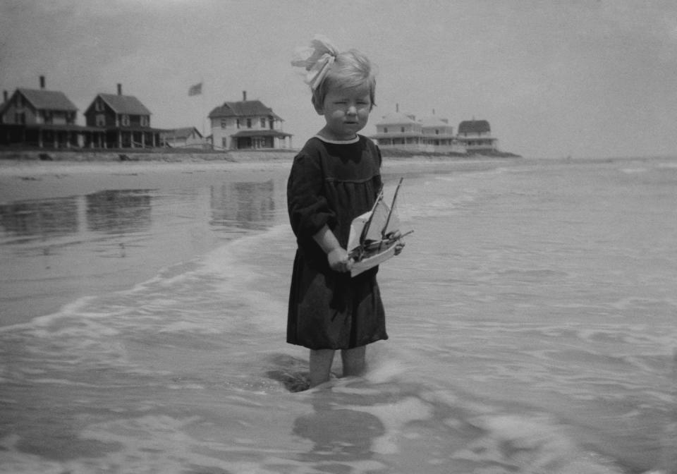 <p>A young girl holding her toy sailboat stands with her feet in the ocean.</p>