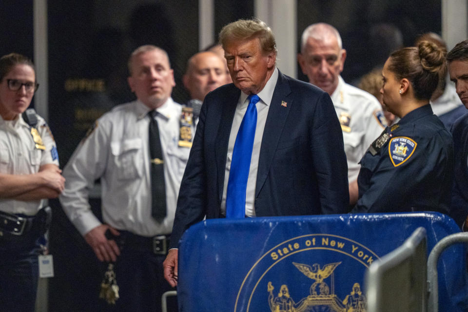 Donald Trump surrounded by several police officers and security personnel with a barrier displaying the State of New York emblem in the foreground