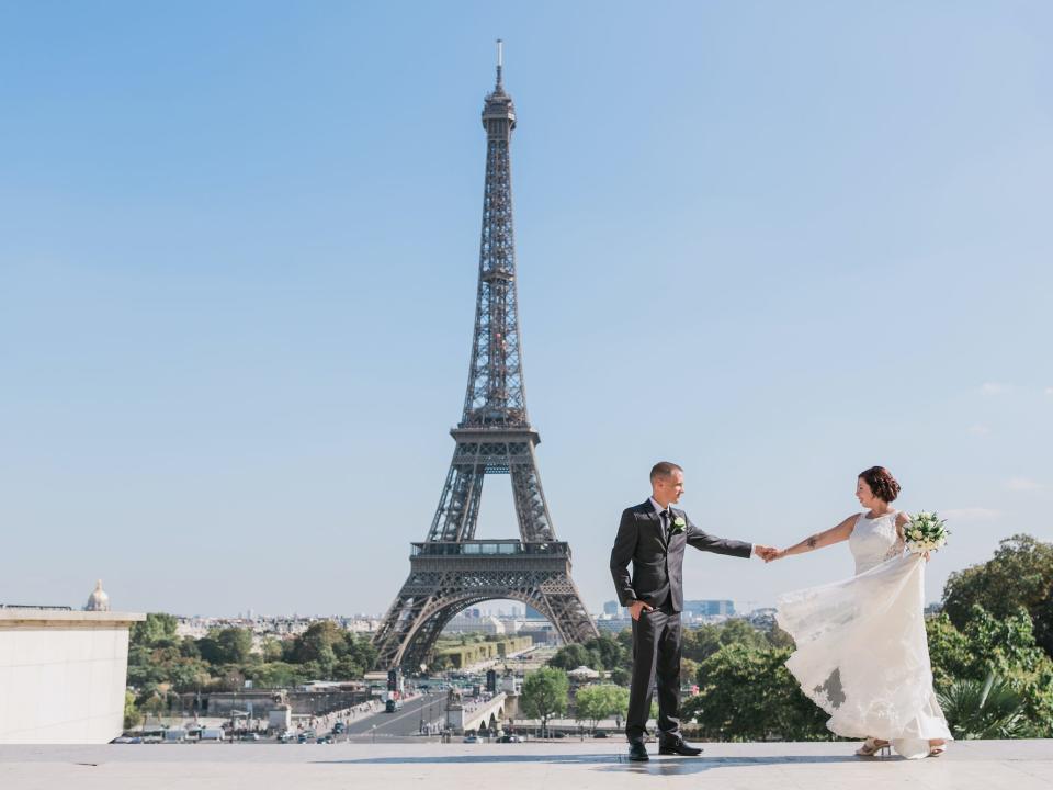 A bride and groom hold hands and look at each other with the Eiffel Tower behind them.