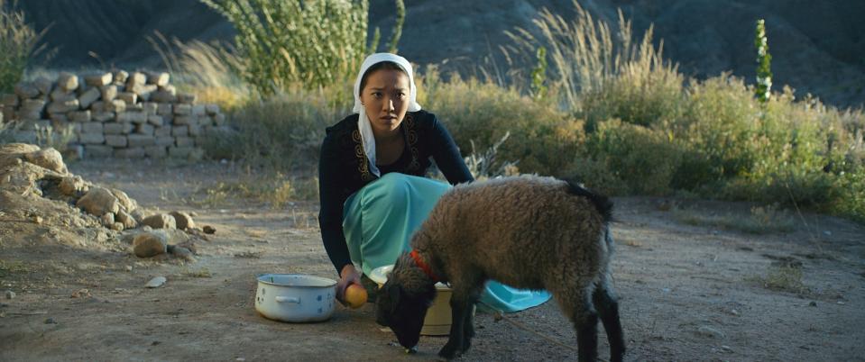 Alina Turdmamatova bent down outside in the dirt with some pots and a goat beside her in a scene from the film