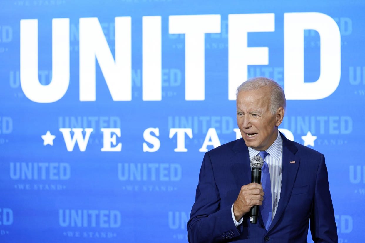 President Biden speaks into a microphone against a blue background displaying the words: United we stand.