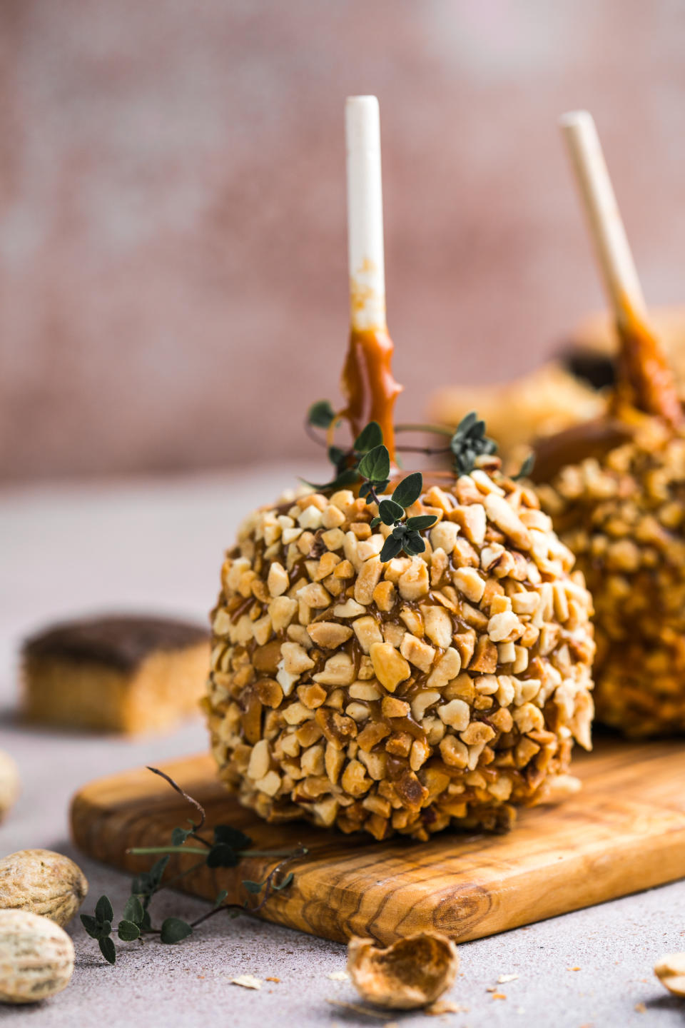 Two caramel apples covered in chopped nuts on a wooden board; blurred background includes dessert items and nuts