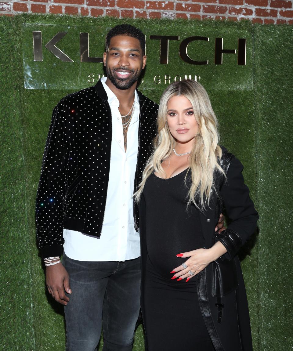 Tristan Thompson and Khloé Kardashian pose together at an event. Thompson wears a studded jacket over a white shirt, and Kardashian wears a black dress with a coat