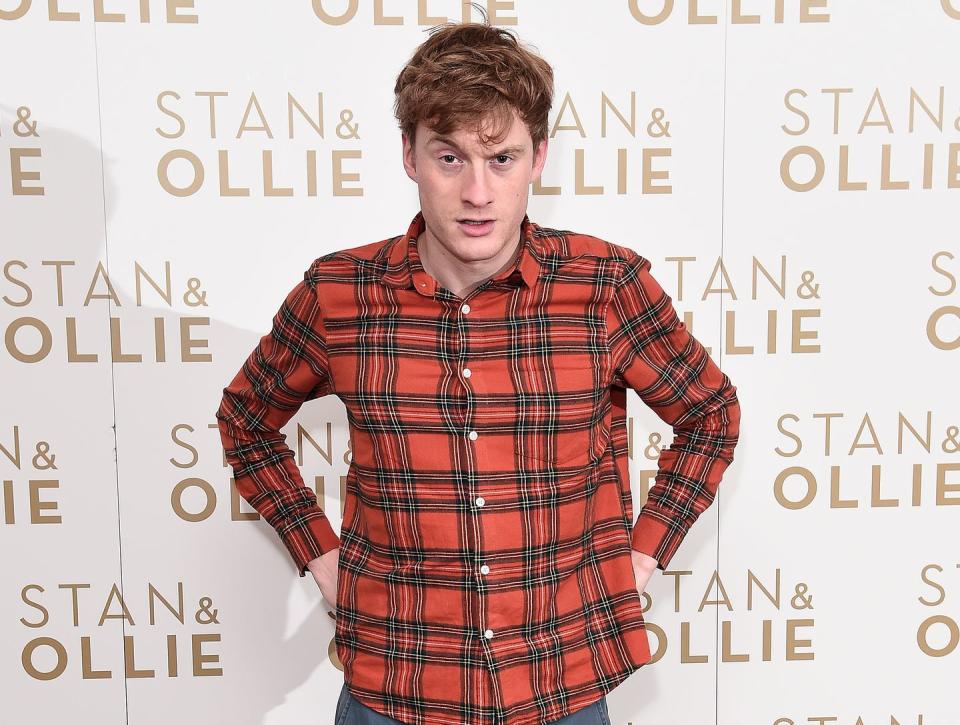 james acaster, a man stands with hands on hips wearing a red check shirt