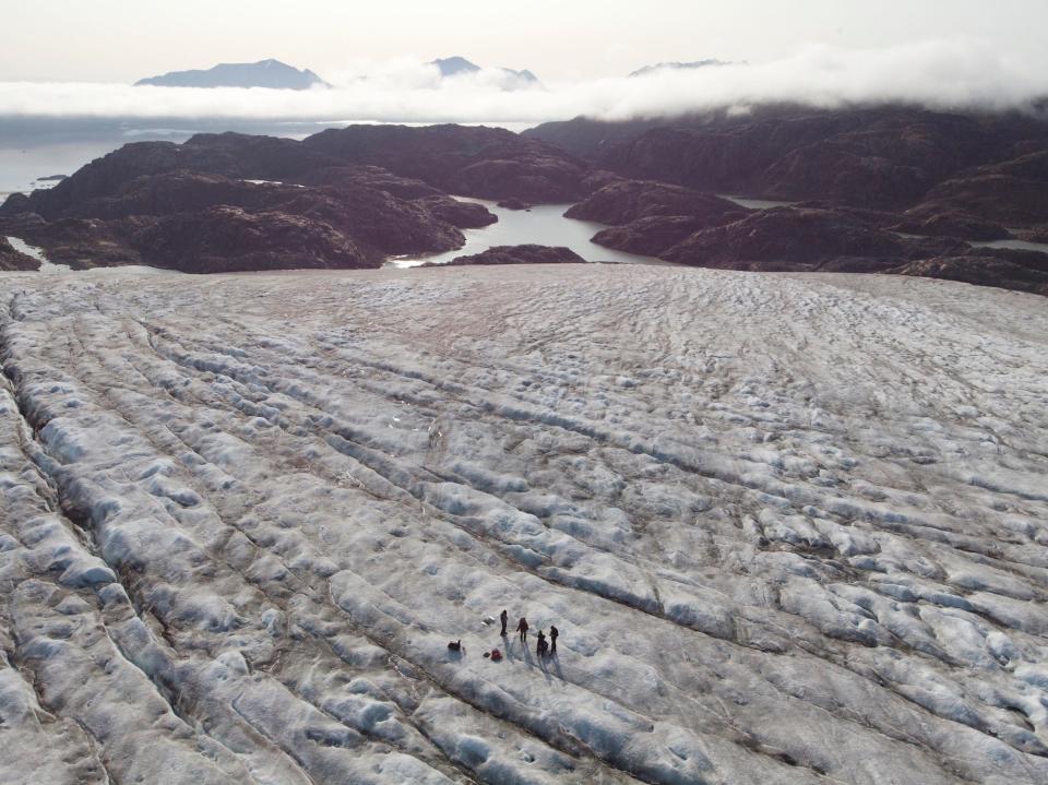 aerial image shows people far below on vast white grey ice field with purple cloud capped mountains in the distance