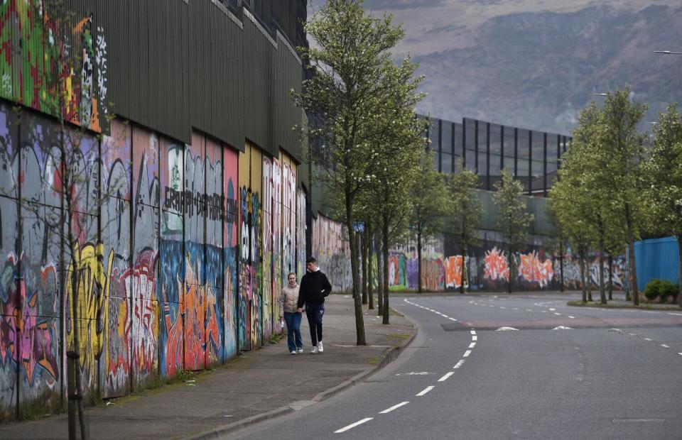 A couple walk along a tall concrete wall covered with graffiti and murals, topped with a wire fence.