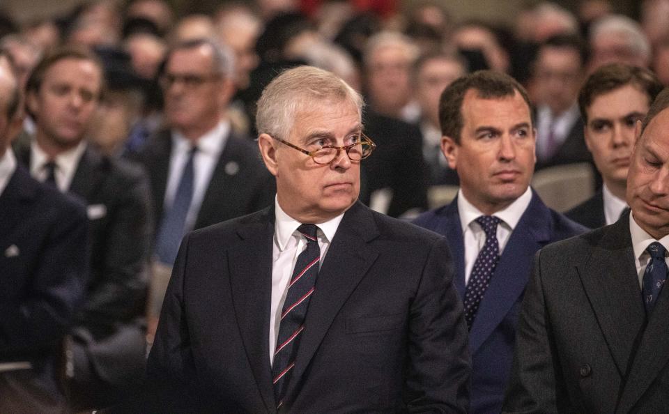 Prince Andrew wears a black suit jacket and stripy tie, he has glasses on and is sitting in Westminster Abbey.