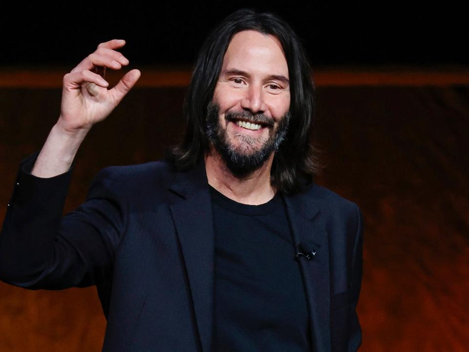 Keanu Reeves, wearing all black, smiles with his hand in the air while onstage at CinemaCon in April 2022.