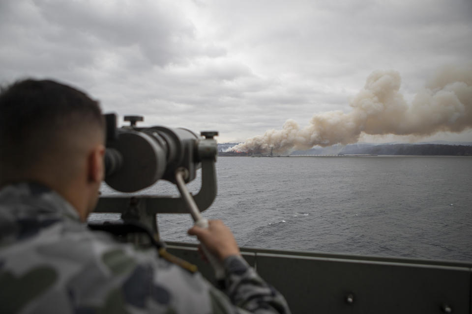 In this photo provided by the Australian Department of Defence on Jan. 6, 2020, Seaman Boatswains Mate Malik El-Leissy watches a burning fire from HMAS Adelaide as the ship arrives at Eden to assist with wildfires. The wildfires have so far scorched an area twice the size of the U.S. state of Maryland. They have destroyed about 2,000 homes. (Able Seaman Thomas Sawtell/ADF via AP)
