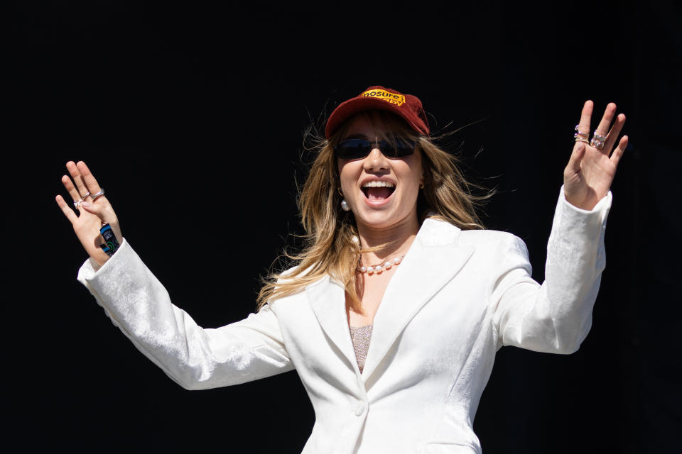 Suki smiling with her arms raised and wearing a cap and suit jacket