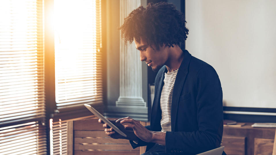 Side view of young handsome African man using his digital tablet while sitting on table at his working place.