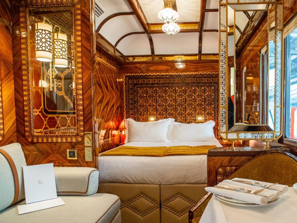 Inside a wood-walled train suite with white and gold furnishings, including a seat on the left, a couch on the right, and a bed in the back center.