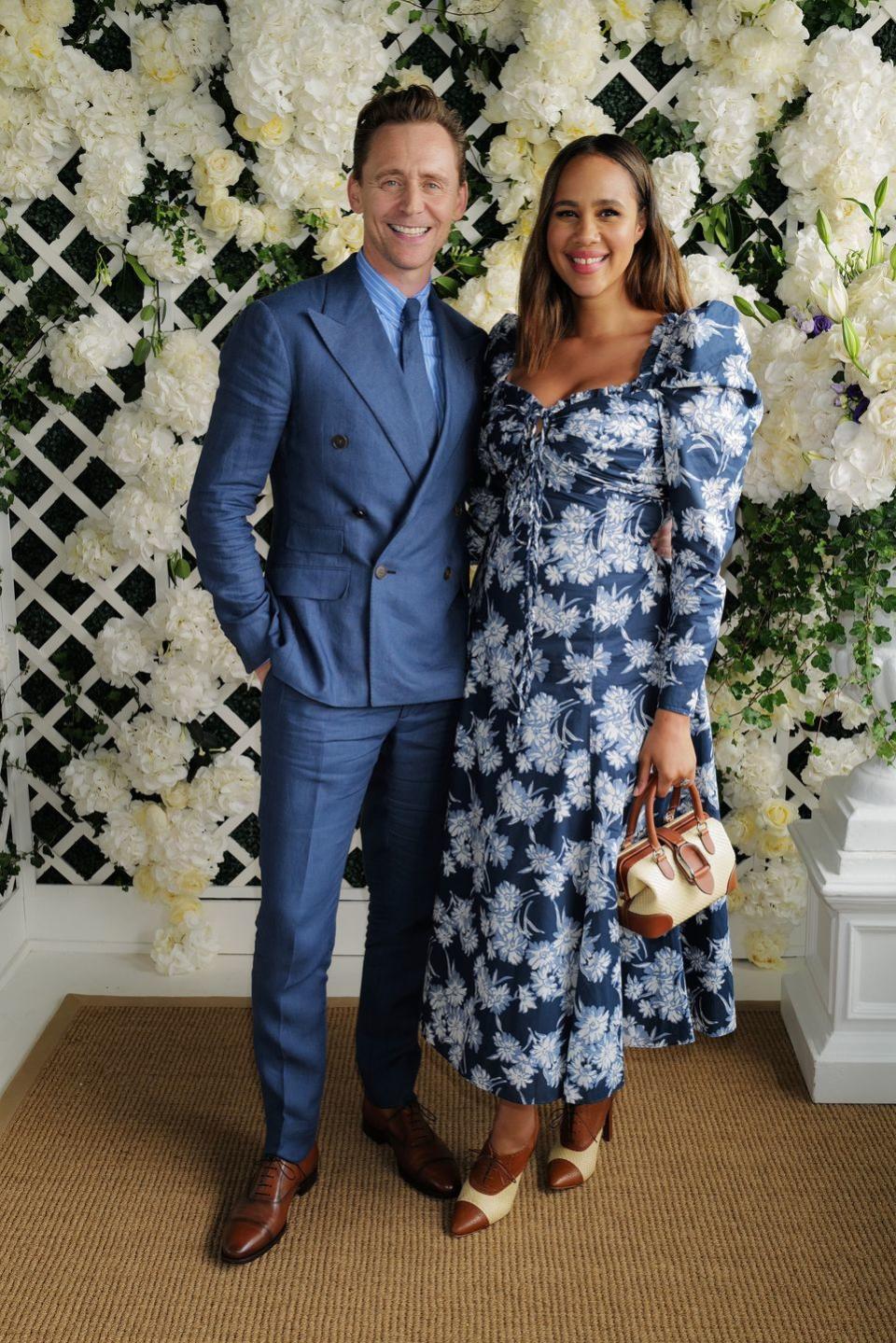 tom hiddleston wearing a blue suit and tie, standing in front of a trellis of white hydrangea flowers next to zawe ashton, who's wearing a long blue and white floral dress, at wimbledon in july 2023