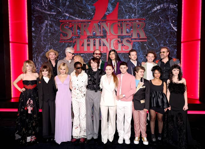 The cast of Stranger Things season 4 smiling at an event, standing in front of backdrop reading "Stranger Things" with a large red number 4 behind it