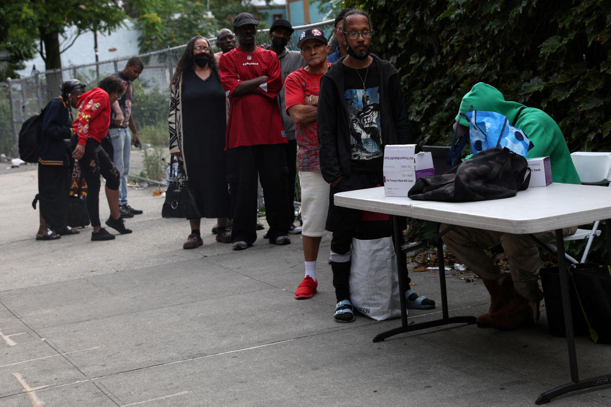 Around 10 people wait in line next to a folding table on a sidewalk, at which someone wearing a green hoodie is hunched over a small sign reading: Viral transport media with swab.