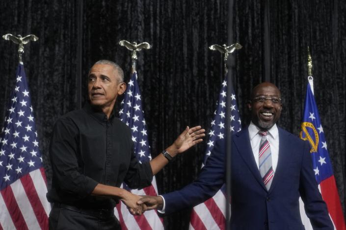 Former President Obama shaking hands with Sen. Raphael Warnock on a flag-filled rally stage.