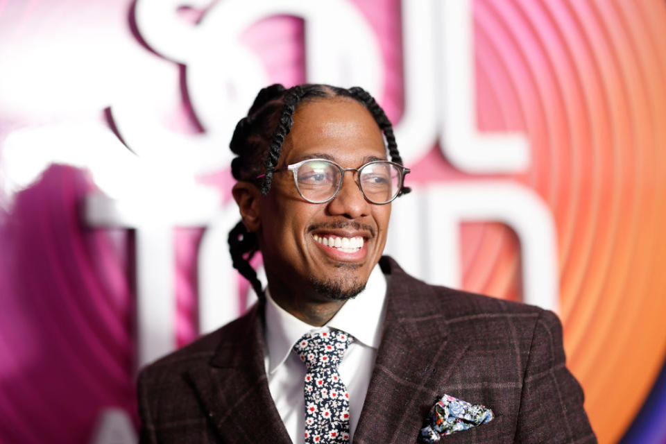 Nick Cannon smiling, wearing glasses, a dark plaid suit, and a floral tie, standing in front of a "Soul Train Awards" backdrop